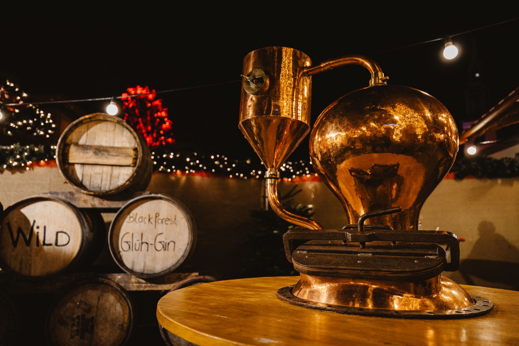 Copper pot still beside small oak barrels under warm string lights