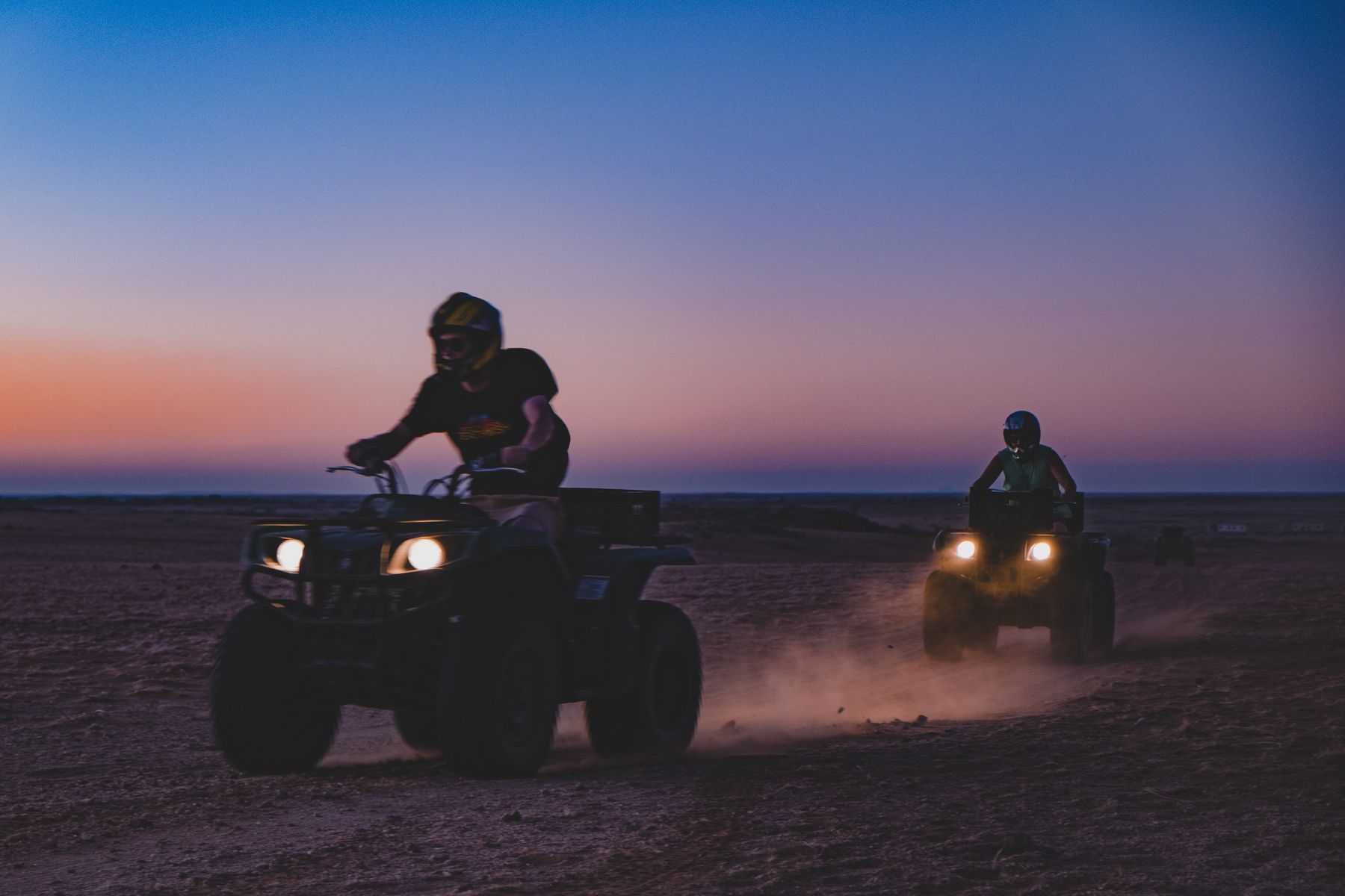 Two riders on quad bikes silhouetted against a purple dusk sky