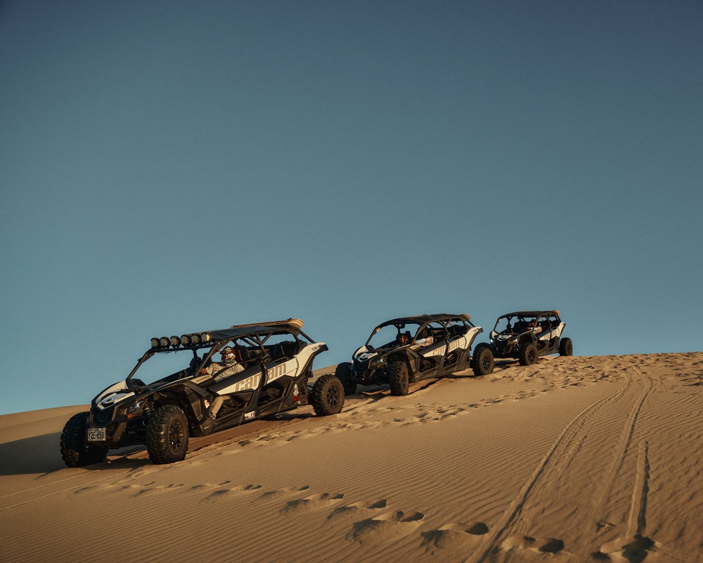 Three side-by-side off-road vehicles parked on a sand dune under a blue sky
